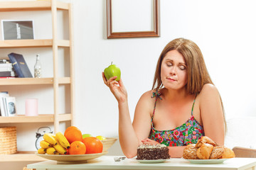 Portrait of beautiful fat woman sitting at table, holding green apple and looking at sweety cake at...