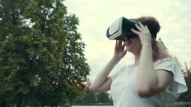 Young woman uses head-mounted display in the park. Playing game using VR-helmet for smart phones.