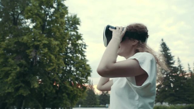 Young woman uses head-mounted display in the park. Playing game using VR-helmet for smart phones.