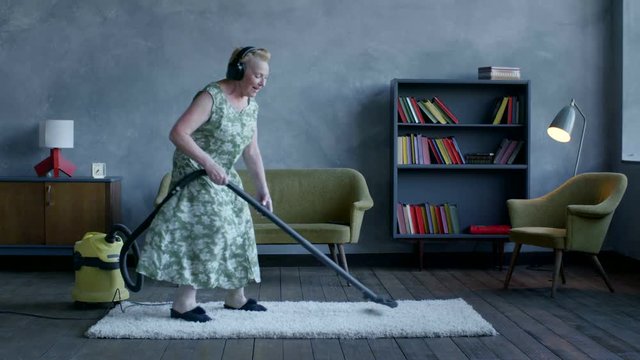 happy elderly woman listening music on headphones and dancing with a vacuum cleaner, home fun