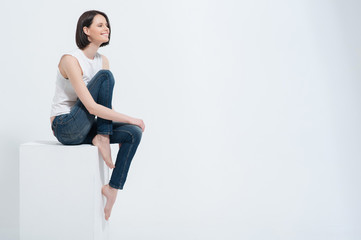 Beautiful young woman sitting on white cube in studio