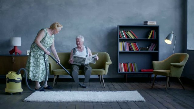 elderly woman vacuuming the floor, and an elderly man reading a newspaper