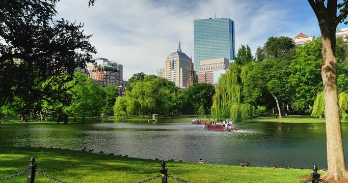 A summer daytime establishing shot of people enjoying the lake in Boston Public Garden. A large rainbow flag is hanging from the John Hancock Building for Gay Pride Month.  	