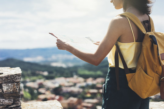 Hipster young girl with bright backpack looking at a map and poining hand the travel plan. View from the back of the tourist traveler on background mountain, sea, binoculars. Mock up for text message