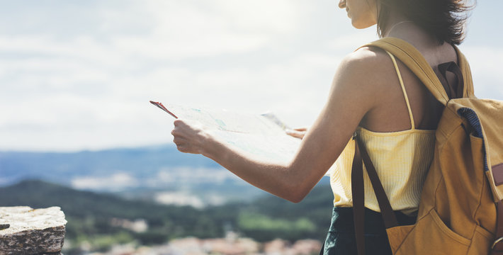 Hipster young girl with bright backpack looking at a map and poining hand the travel plan. View from the back of the tourist traveler on background mountain, sea. Mock up for text message