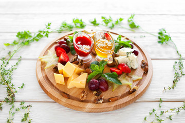 Plate with snacks for wine in herbal frame on white. Top view on few kinds of cheese and fruits on catering platter with red and yellow sauces. Wine snacks plate on white wooden background