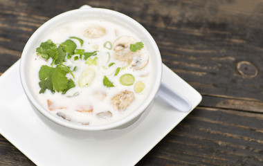 Thai soup in a white dish on a wooden table. Spicy coconut milk soup with shrimp and greens. 