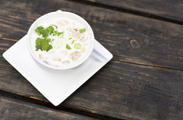 Thai soup in a white dish on a wooden table. Spicy coconut milk soup with shrimp and greens. 