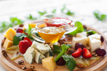 Wine snacks set on wooden cutting board closeup. Fruits and different kinds of cheese with yellow and red sauces on round wooden board. Strawberry, grape, nuts and cheese