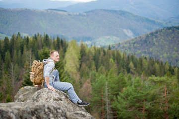 Courageous tourist male with a backpack sitting on the top of a rock, with an astonishing blurred view on forest valley and hills