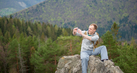 Naklejka na ściany i meble Portrait of young climber sitting on the peak of rock makes the photo on the his phone on the blurred background of forest valley