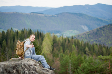 Naklejka na ściany i meble Smiling tourist male with a backpack sitting on the edge of a rock and showing thumb up gesture of good class, with an astonishing blurred view on forest valley and hills