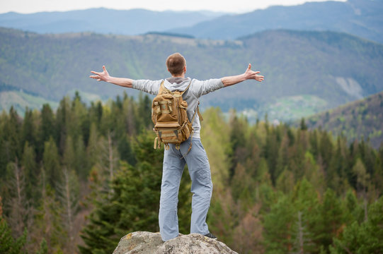 Male climber with brown backpack standing on the top of a rock with back to the camera, his arms out to the sides and enjoying picturesque view of on the green forest and nice mountains