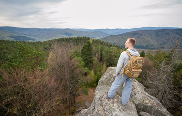 Courageous man with a backpack standing on the edge of a rock and looking into the distance on the green forest and nice mountains