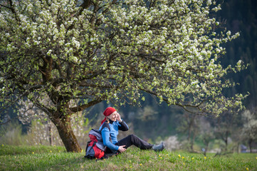 Fototapeta na wymiar Woman with a backpack enjoy under blooming tree on top of hill with yellow wildflowers, green grass and talking on the mobile phone with forest valleys as blurred background.