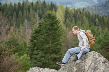 Naklejka premium Man with a brown backpack sitting on the peak of rock and looking down on the blurred background of forest valley and hills. Spring nature.