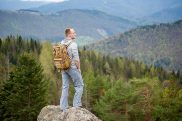 Cool man with a backpack standing on the edge of a rock and looking into the distance on the green forest and nice mountains.