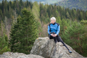 Naklejka na ściany i meble Athletic old female in a blue jacket with climbing equipment sitting on the top of the rock on the blurred background of forest valley.