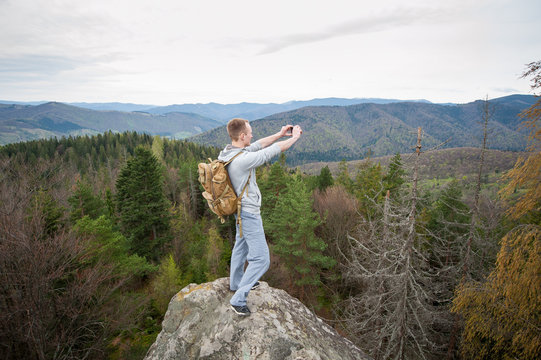 Brave male climber with brown backpack standing on the peak of a rock, with back to the camera and makes the photo on the his phone with forest valley and nice hills.