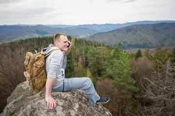 Young brave man with a backpack sitting on the peak of rock, looking away and talking on the phone on the background of forest valley and hills.