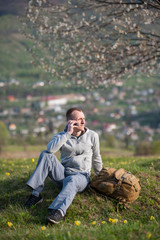 Naklejka na ściany i meble Young man with backpack sitting on hill with green grass yellow wildflowers and talking on the mobile phone on the blurred background of blooming tree and small town in the distance