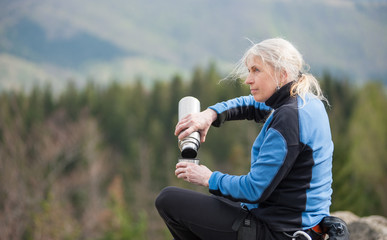 Adult female climber in a blue jacket sit on the peak of rock and poured from a thermos against forest valley with blurred background. Close-up