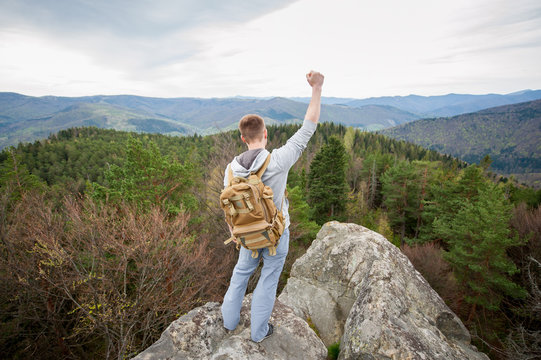 Tourist with a brown backpack standing on the top of the rock, back to the camera, with a hand up, against natural landscape of green forest and beautifull hills. Wide angle lens