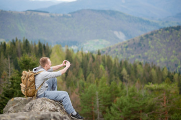 Fototapeta na wymiar Successful person with backpack sitting on the rock makes the photo on the his phone with forest and mountains in blurred background