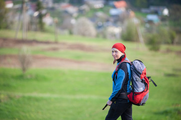 Fototapeta na wymiar Traveler woman in a red scarf on her head and blue jacket with a backpack looking at the camera, blurred greenery background. Close-up