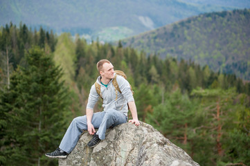 Naklejka na ściany i meble Rock climber with a brown backpack sitting on the peak of rock and looking into the distance on the blurred background of forest valley and hills. Spring nature.