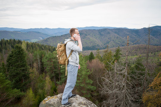 Tourist with brown backpack standing on the peak of a rock, with back to the camera, talking on the phone and looking into the distance on the green forest and nice mountains.