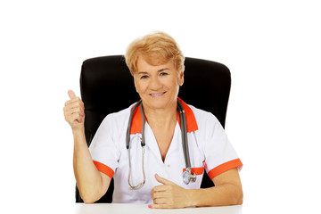 Smile elderly female doctor or nurse sitting behind the desk and shows thumb up