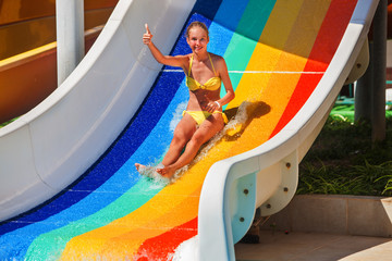 Child teenager wearing bikini slides on water slide and shows thumb up at aquapark . Slide painted...