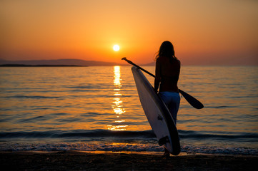 Junge Frau mit SUP Paddelboard geht in den Sonnenuntergang