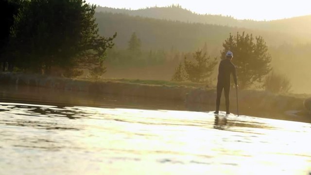From a wide shot with a slightly canted angle, a man in a wetsuit paddleboards down an Oregon river away from camera during a beautiful, glorius sunset
