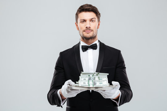 Attractive butler in tuxedo and gloves holding money on tray