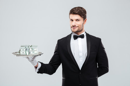 Gorgeous butler in tuxedo standing and holding tray with money