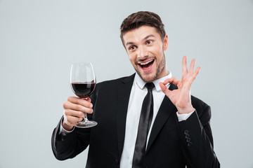 Cheerful drunk businessman drinking red wine and showing ok sign