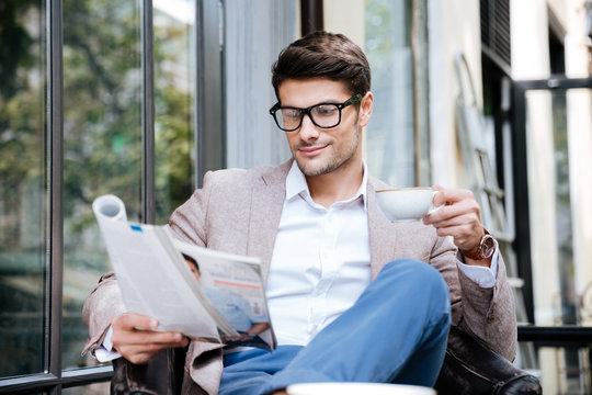 Handsome young man with magazine drinking coffee in outdoor cafe - Powered by Adobe