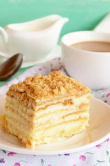 A piece of Napoleon cake. Cake of puff pastry with sweet cream on a plate. A cup of coffee milk. Spoon, fabric with floral pattern. Sweet fresh dessert. Home baking. Close-up