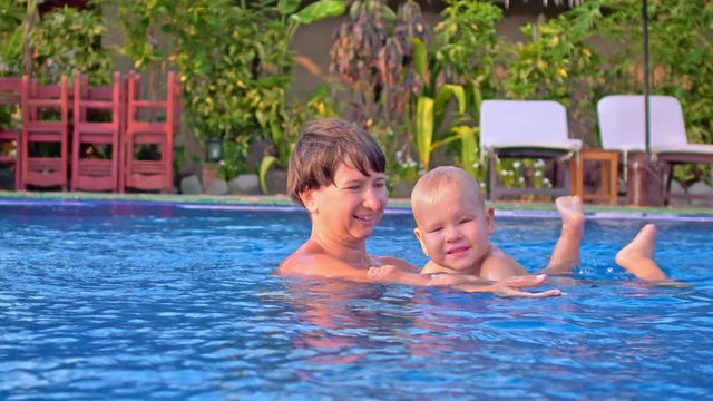 Toddler boy tries to swim in outdoor pool while his mother holds and supports him. Tropical resort in background.
