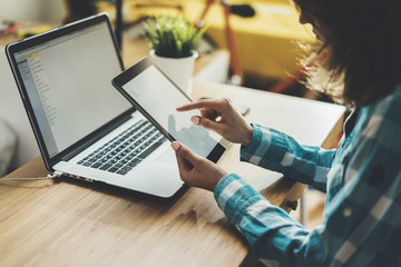 Close-up of young hipster girl sitting at workspace and using modern digital tablet and laptop,...