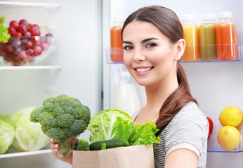 Young woman with purchase box full of vegetables standing beside fridge