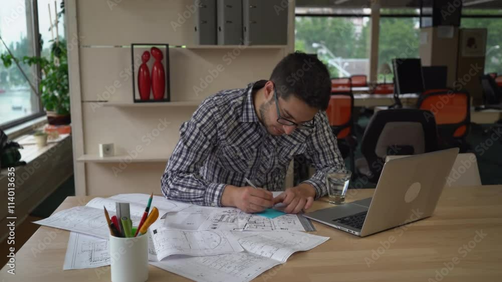 Aufkleber Architect checking details and make notes on the paper plan. Documents and computer on the wooden table with equipment for drawing and draftsmanship. Male wearing in casual clothes work in modern