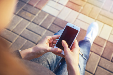 Close up of a woman's hands using white smartphone with blank screen, sitting on a bench in the park. Style of casual, sunny day, sneakers, photo filter.