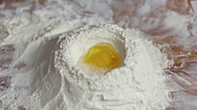 Close up pouring raw egg into well of flour on wooden table