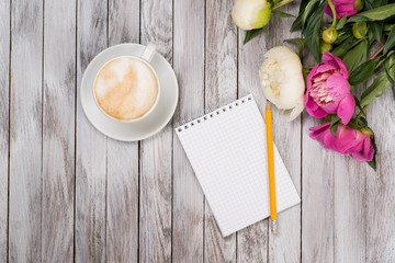Notebook with a pencil next to coffee and peonies flowers on wooden background. Top view.