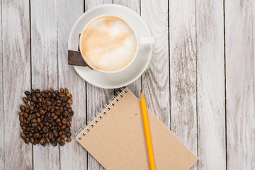 Notebook with a pencil next to coffee and coffee beans, piece of chocolate on white wooden background. Top view.