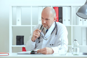 Old male medicine doctor  sitting at the table and holding glasses reading something in diary. Medical care or insurance concept.