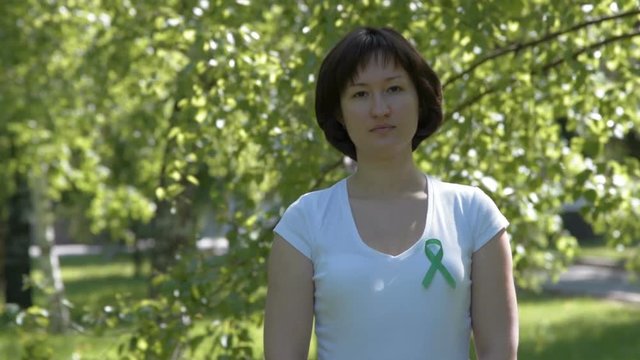 Young brunnete woman in white t-shirt wearing green awareness ribbon, trees background. Kidney cancer, glaucoma, organ donation, depression, environmental protection awareness